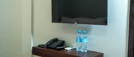 A wall-mounted TV above a wooden shelf with a phone, remote controls, and two water bottles.
