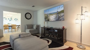 A modern living room with a sofa, a dining area with chairs, a large painting, a clock, and a floor lamp.