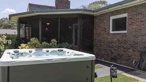 A hot tub in a backyard with a brick house, a covered patio, and a garden.