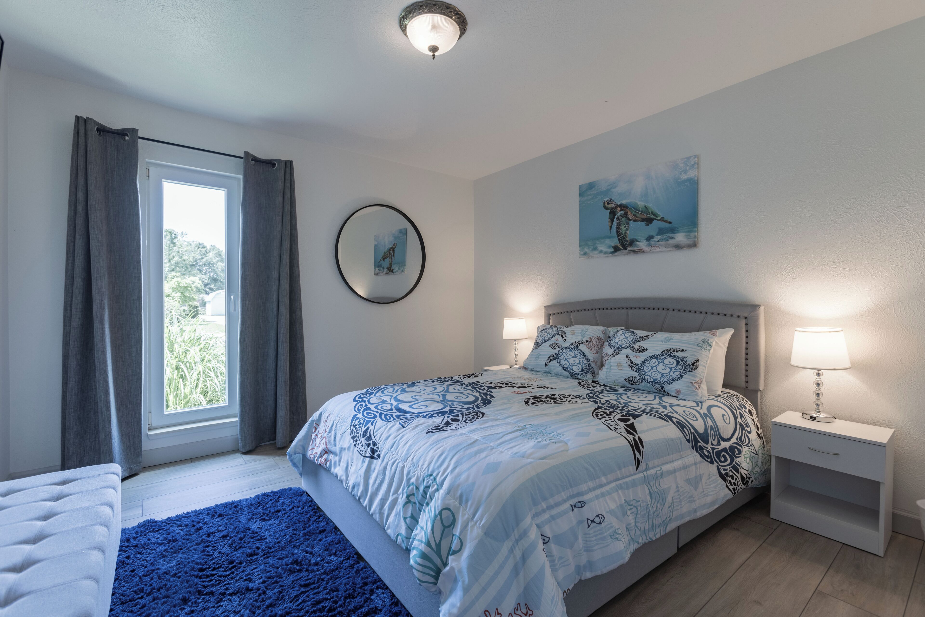 A bedroom with a bed, bedside tables, a window with curtains, a wall-mounted mirror, and a painting.