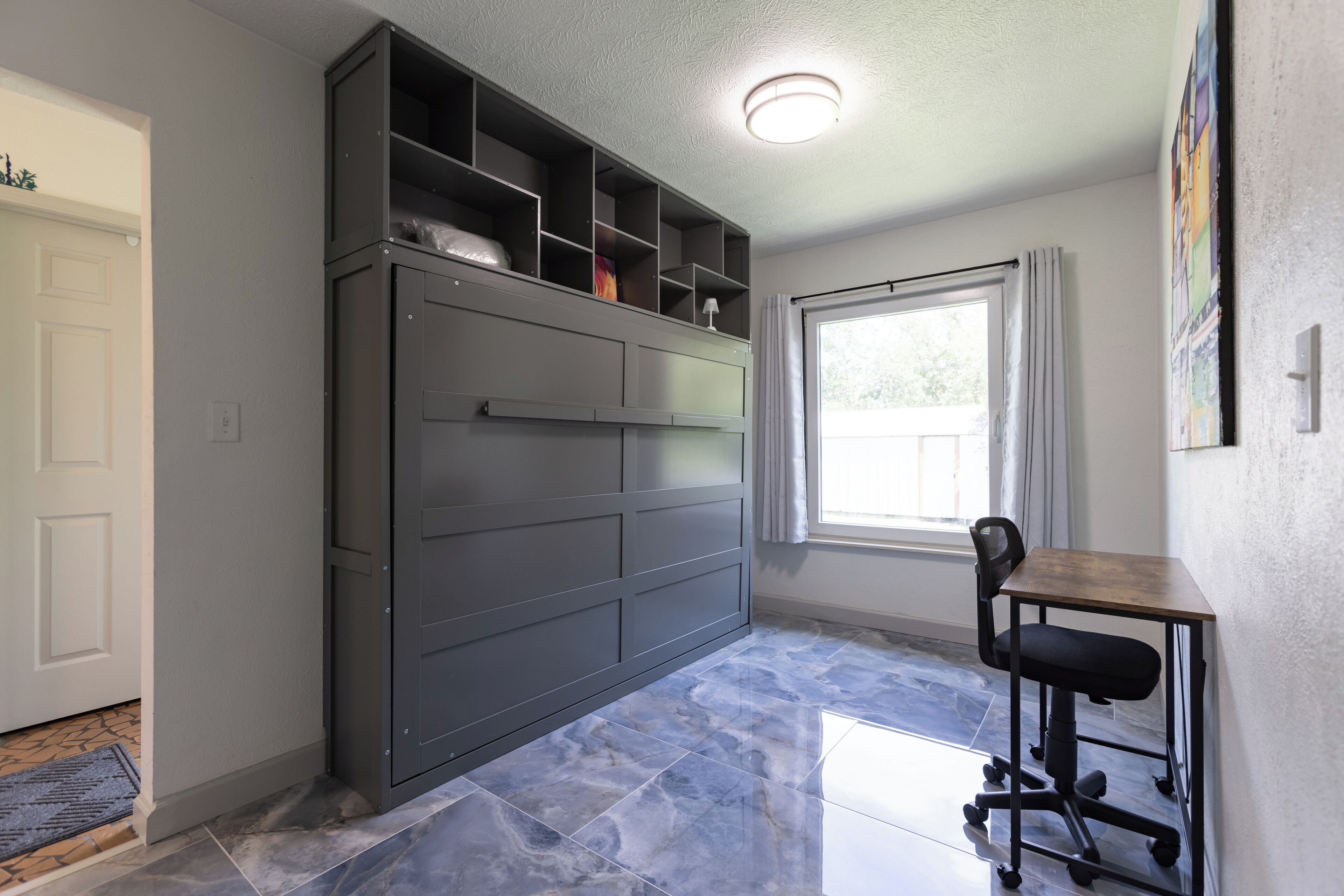 A room with a built-in desk and chair, a window with curtains, and a tiled floor.