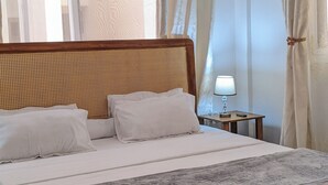 Un letto con testiera in vimini, un comodino con una lampada e tende leggere.