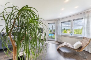 Un salón moderno con una planta grande, una silla de mimbre, un sofá y vista a una piscina a través de un ventanal.