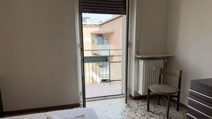 Una camera d'albergo con un letto, una sedia, un termosifone e vista su un balcone e un edificio.