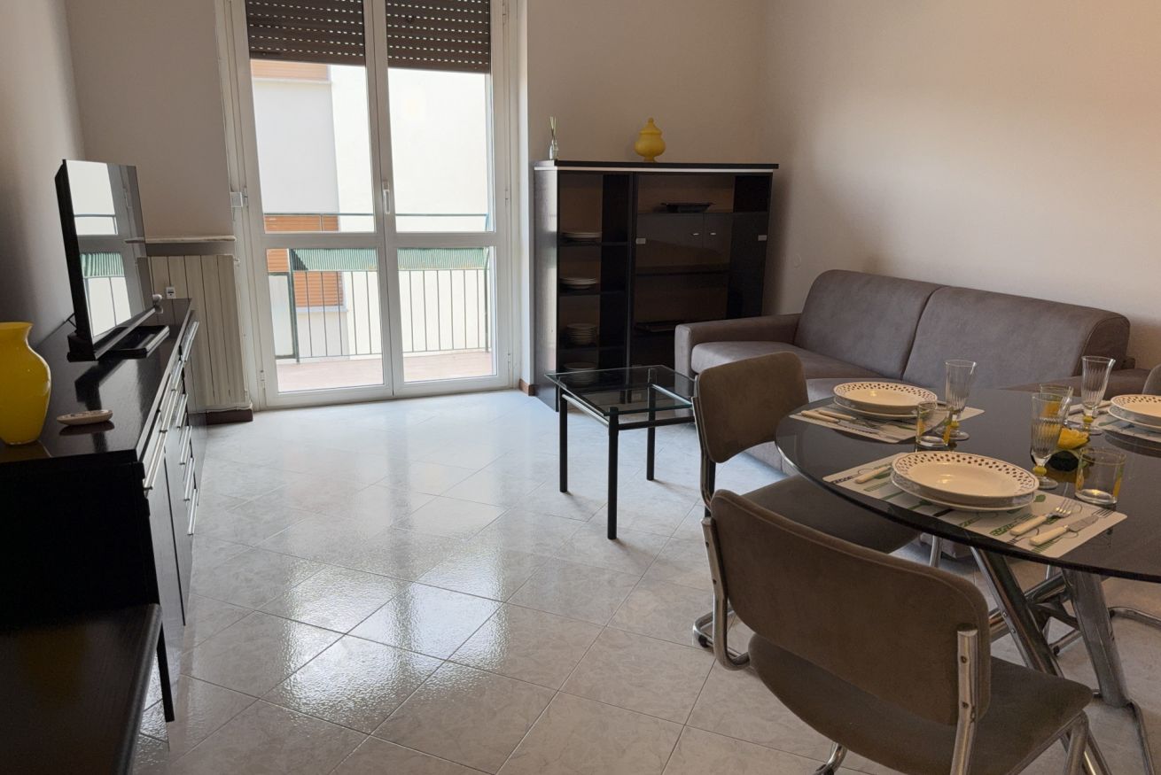 Ein Essbereich mit einem Glastisch, gedeckt für vier Personen, einem grauen Sofa und einer Glastür zum Balkon.