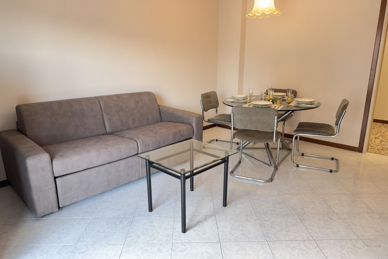 A modern living room with a grey sofa, a glass-top coffee table, and a dining table with four chairs.