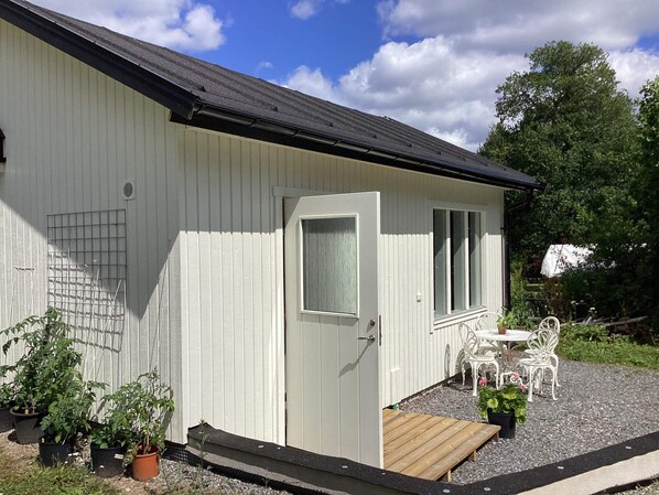 A small, single-story building with a white exterior, a black roof, and a door with a window. There is a small porch area with a table and chairs, and several potted plants are visible. - 4 Person Holiday Home in Vaxholm (Vaxholm)