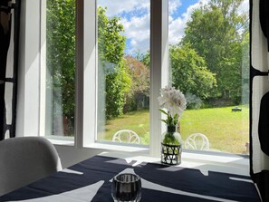 Ett rum med ett bord dukat för middag, stora fönster med utsikt över en trädgård och en vas med blommor på fönsterbrädan.