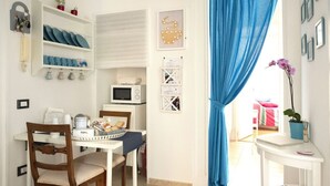 A dining area with a white table, chairs, and a microwave. There are shelves with plates and a potted plant. - At Giulia's Home Vomero Apt (NAPOLI)