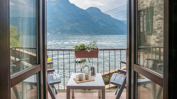 A balcony with a table, chairs, a hanging plant, and a view of a lake and mountains.