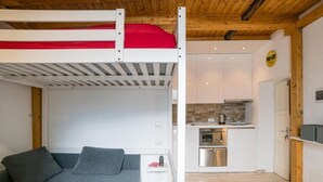 A bunk bed with a red mattress, a gray sofa, a kitchen with modern appliances, and a wooden ceiling.