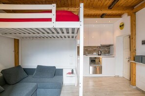 Un lit superposé avec un matelas rouge, un canapé gris, une cuisine équipée d’appareils modernes et un plafond en bois.
