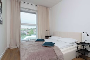 Une chambre avec une grande fenêtre, un lit avec deux oreillers, une table de chevet avec une lampe, et une vue sur le paysage urbain.