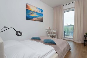 A bedroom with a bed, a painting of a sunset over the ocean, a window with curtains, and a balcony with a chair.