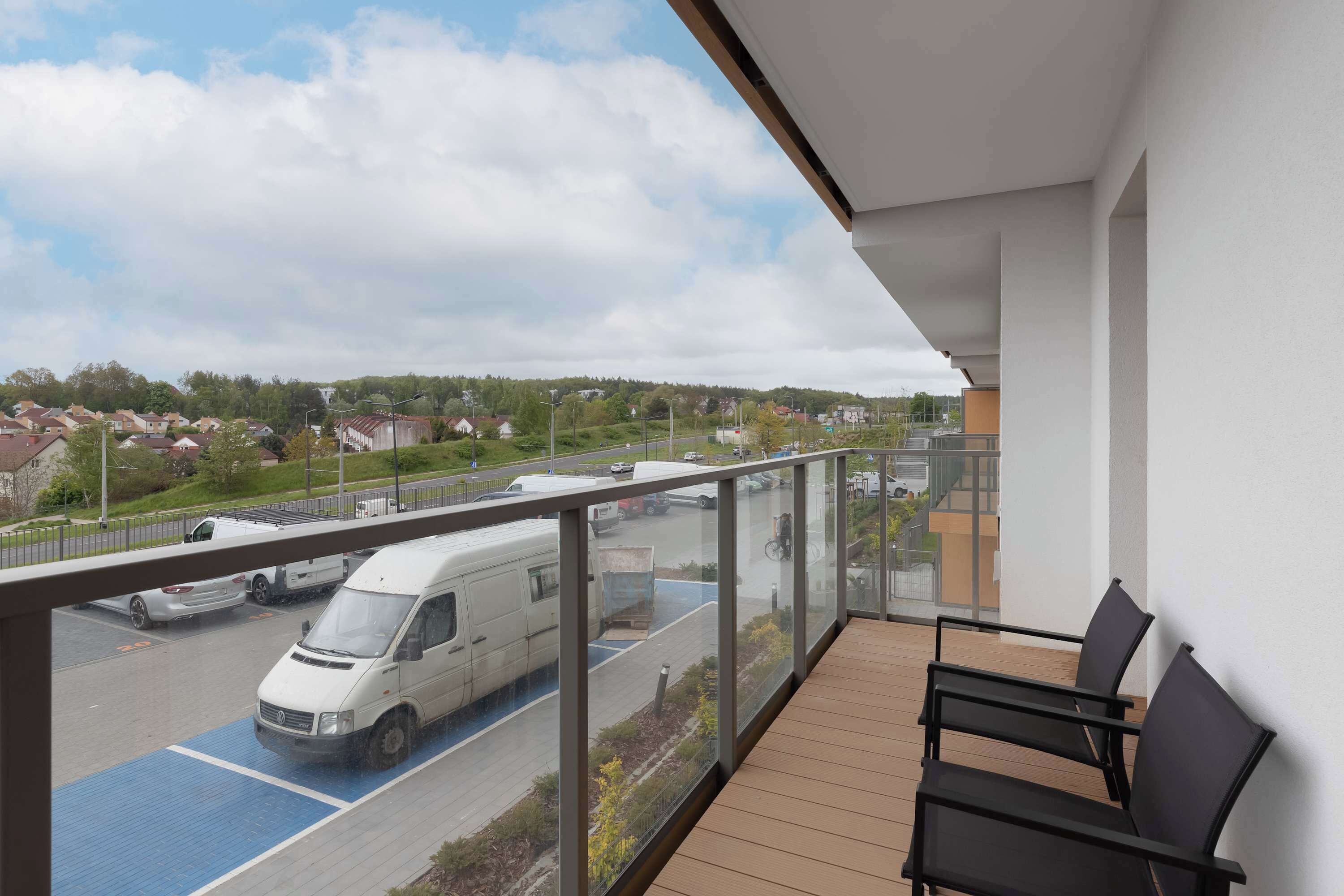 Un balcon donnant sur un parking et un fourgon, avec un plancher en bois et des chaises noires.