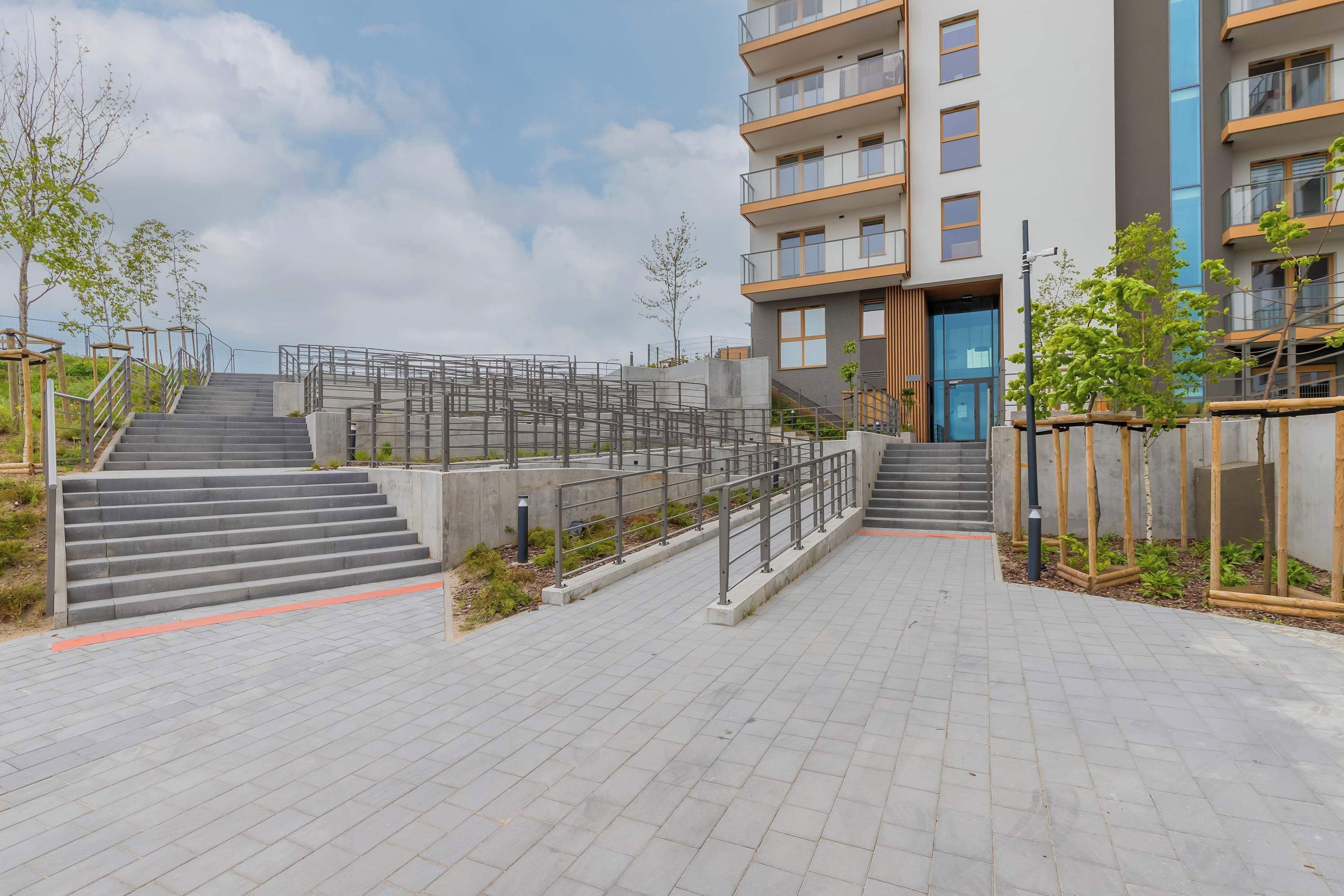 Een modern gebouw met een trap die naar de ingang leidt, omgeven door een verhard gebied met leuningen en begroeiing.