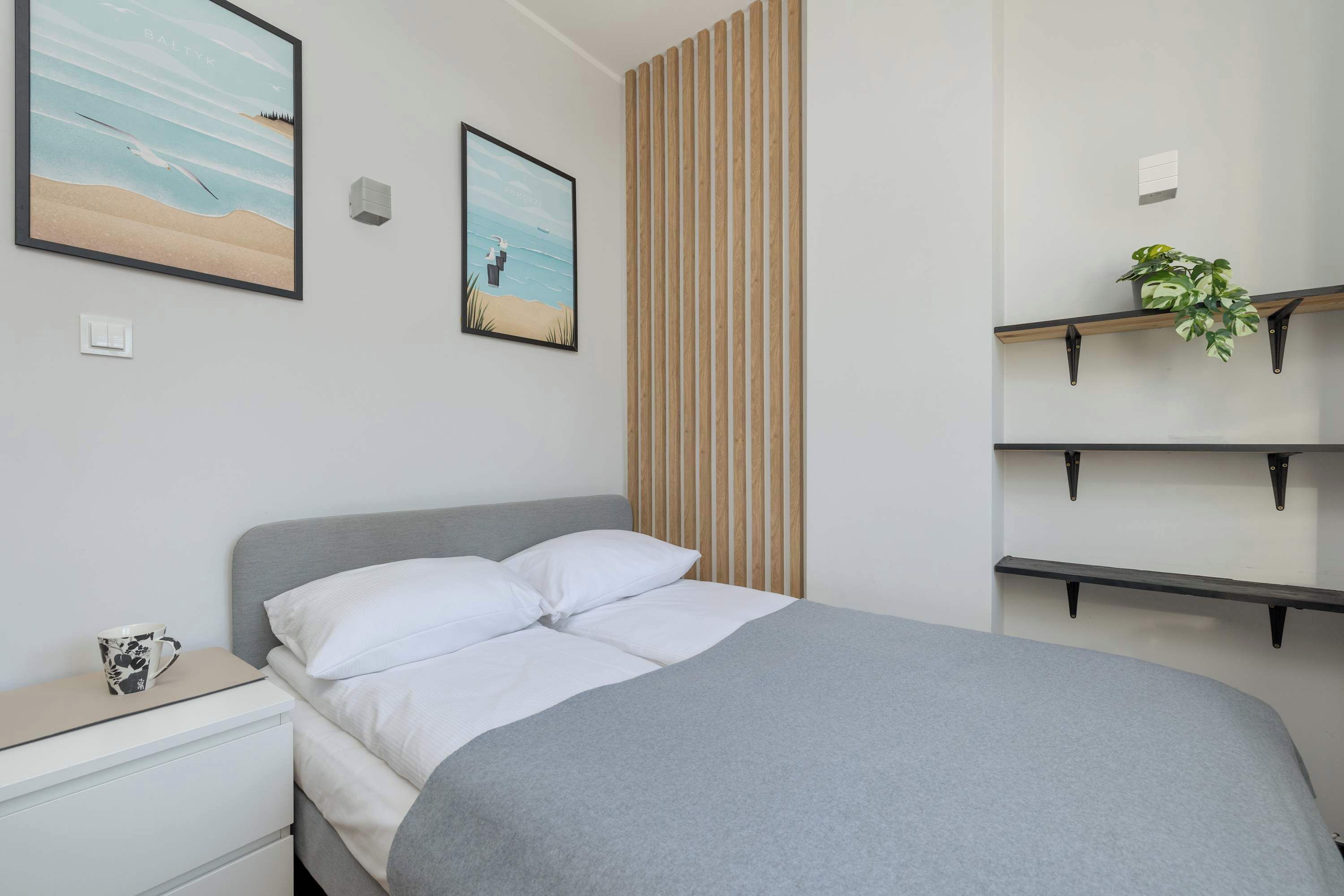 Una cama bien hecha con ropa de cama blanca, una mesita de noche con una taza y dos cuadros en la pared.