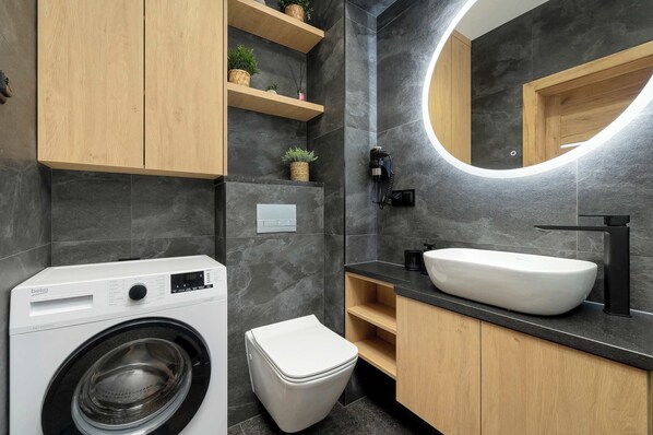 Un baño moderno con lavadora, inodoro, lavamanos y estanterías de madera con plantas.