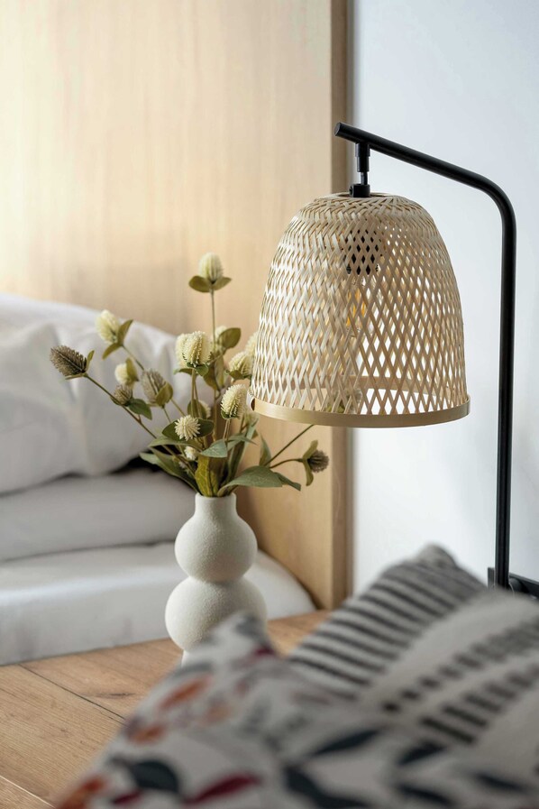 Una habitación con una cama, un jarrón blanco lleno de flores y una lámpara con pantalla tejida.