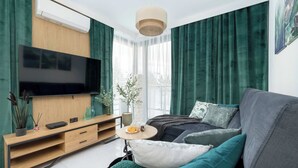 Un soggiorno moderno con una televisione a schermo piatto, un mobile TV in legno, tende verdi, un divano grigio e un tavolino rotondo con una pianta e un teiera.