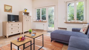 A living room with a sofa, a TV, a wooden cabinet, a table with fruit, and a view of greenery through the windows. - Family Apartment with Terrace | 2 km to Krupowki and Parking (Zakopane)