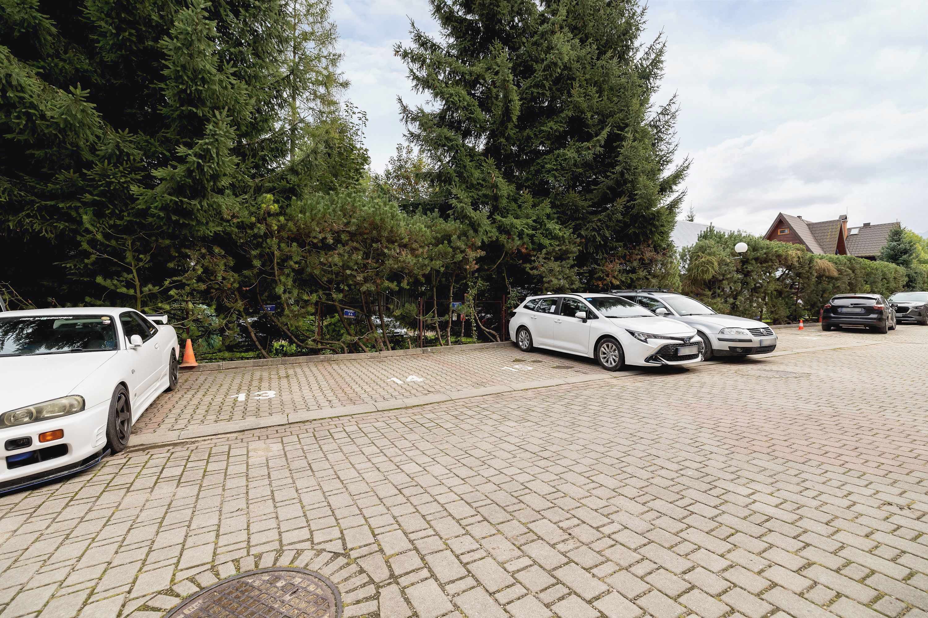 Ein Parkplatz mit mehreren Autos, darunter ein weißer Sportwagen und eine weiße Limousine, umgeben von Bäumen und einem Gebäude im Hintergrund.