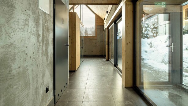 Un pasillo con techo y paredes de madera, una puerta de cristal que da al exterior, y vistas de un paisaje nevado.