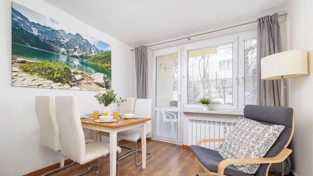 A dining area with a table and chairs, a window with curtains, a lamp, and a painting of a mountain landscape.