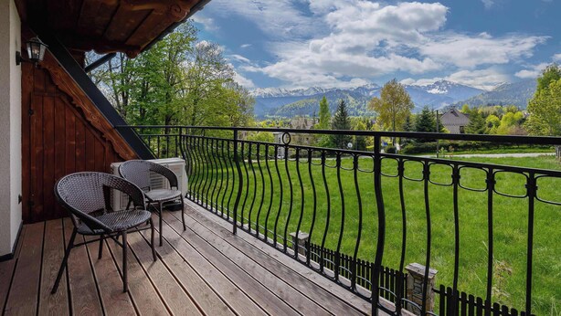A wooden deck with two wicker chairs and a table, overlooking a green lawn and mountains.