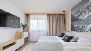 Een moderne slaapkamer met een bed, een aan de muur bevestigde televisie, een groot raam met gordijnen en een schilderij.