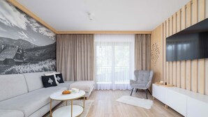 Een moderne woonkamer met een bank, een salontafel, een aan de muur bevestigde televisie en een groot raam met gordijnen.