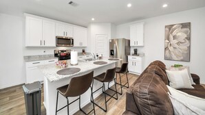A modern kitchen with white cabinets, stainless steel appliances, and a central island with bar stools. - Marice Serene Home at Katy Fulshear  (Fulshear)
