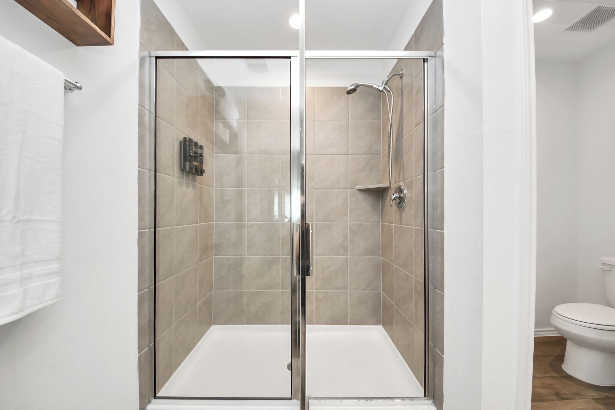 A glass-enclosed shower stall with a white toilet and a wooden shelf above it.
