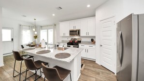A modern kitchen with white cabinets, a stainless steel refrigerator, and a central island with bar stools. - Marice Serene Home at Katy Fulshear  (Fulshear)