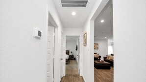 A hallway with white walls, wooden flooring, and a door leading to a living area with a sofa and a coffee table. - Marice Serene Home at Katy Fulshear  (Fulshear)