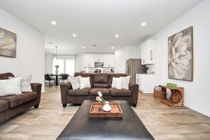 A modern living room with a brown leather sofa, a coffee table with a vase, and a dining area in the background. - Marice Serene Home at Katy Fulshear  (Fulshear)