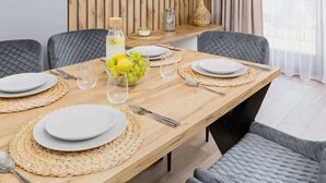 Un espace repas moderne avec une table en bois, des chaises à revêtement gris, un téléviseur fixé au mur et un vase contenant des plumes d’herbe des pampas.