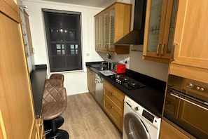 A compact kitchen with wooden cabinets, a built-in oven, a washing machine, and a window with blinds.