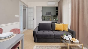 A modern living room with a grey sofa, a small coffee table, and a dining table with plates and cutlery. A kitchen is visible in the background with a microwave and cabinets. - Stylish Apartment in Mokotow | Air Conditioning and Parking (Warszawa)