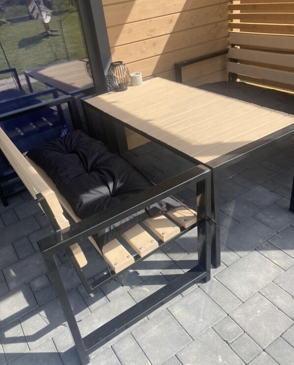 A convertible sofa bed with a glass table and metal frame, set on a paved patio with a wooden wall and a garden view. - Kalimera domki Rewal  (Rewal)