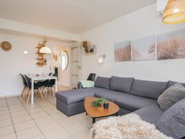 Una sala moderna con un sofá gris, una mesa de centro redonda con una planta, una zona de comedor con una mesa y sillas blancas, y un espejo grande en la pared.