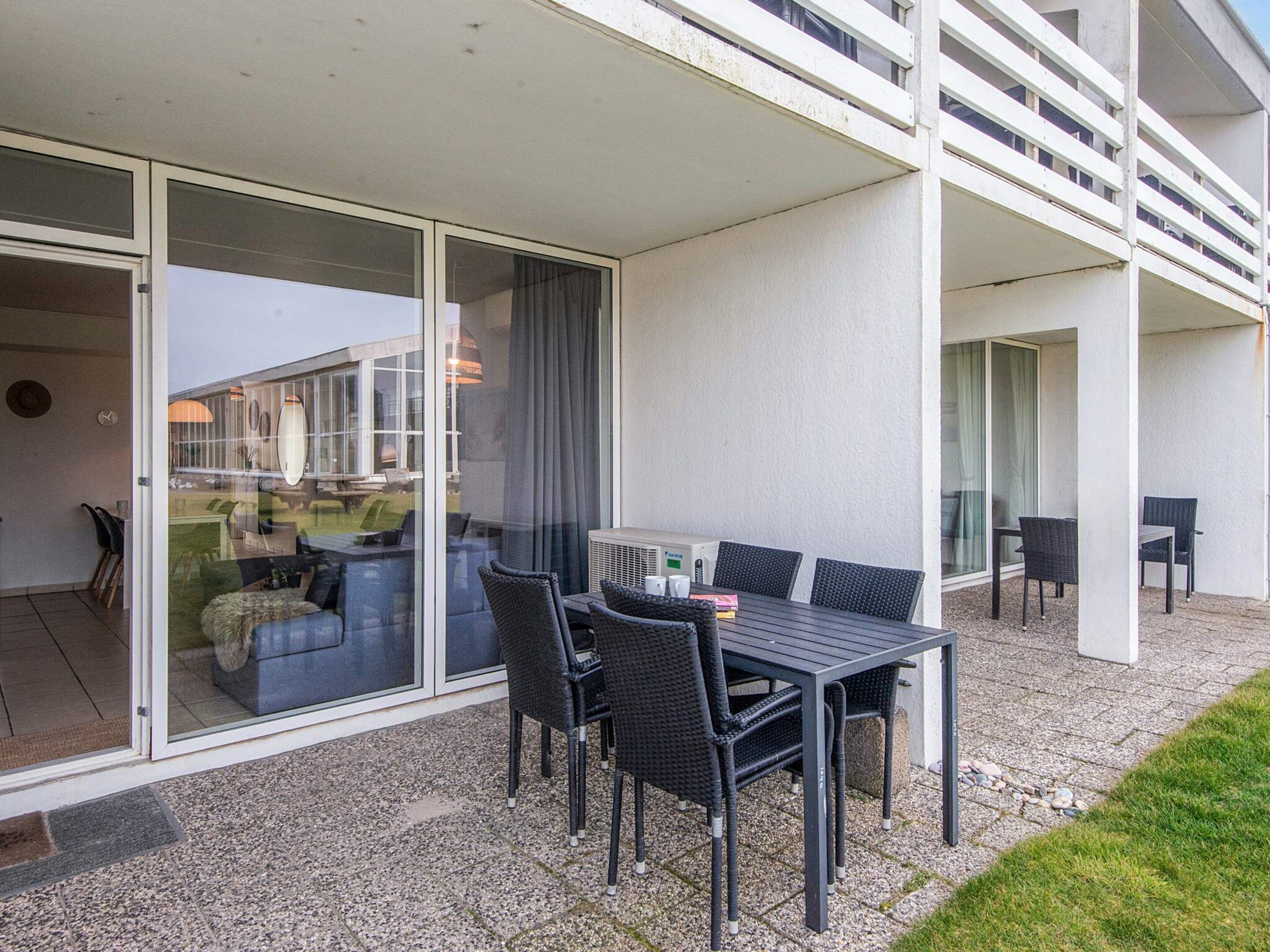 A modern outdoor dining area with a table and chairs, a sliding glass door leading to an interior space, and a building with balconies.