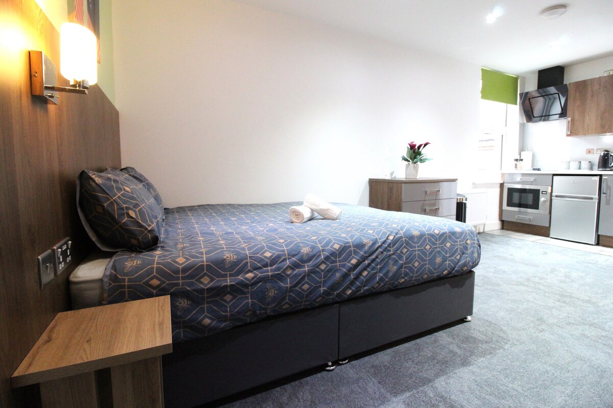 A modern hotel room with a bed, bedside table, and a kitchenette area.
