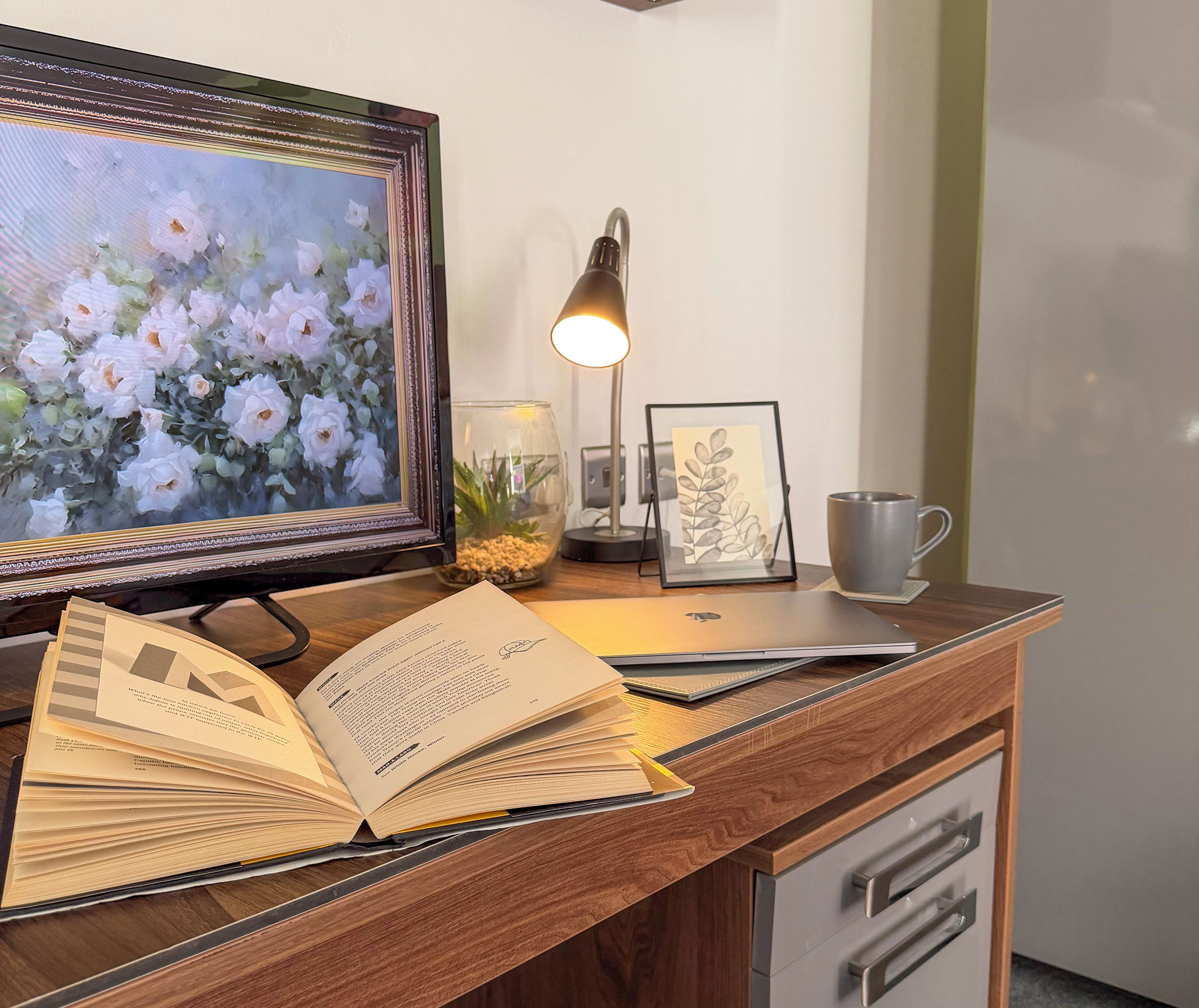 Een houten bureau met een laptop, een open boek, een lijst, een bureaulamp en een mok.