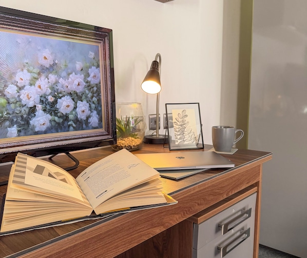 A wooden desk with a laptop, an open book, a framed painting, a desk lamp, and a mug. - Victoria House by CovStays (Coventry)