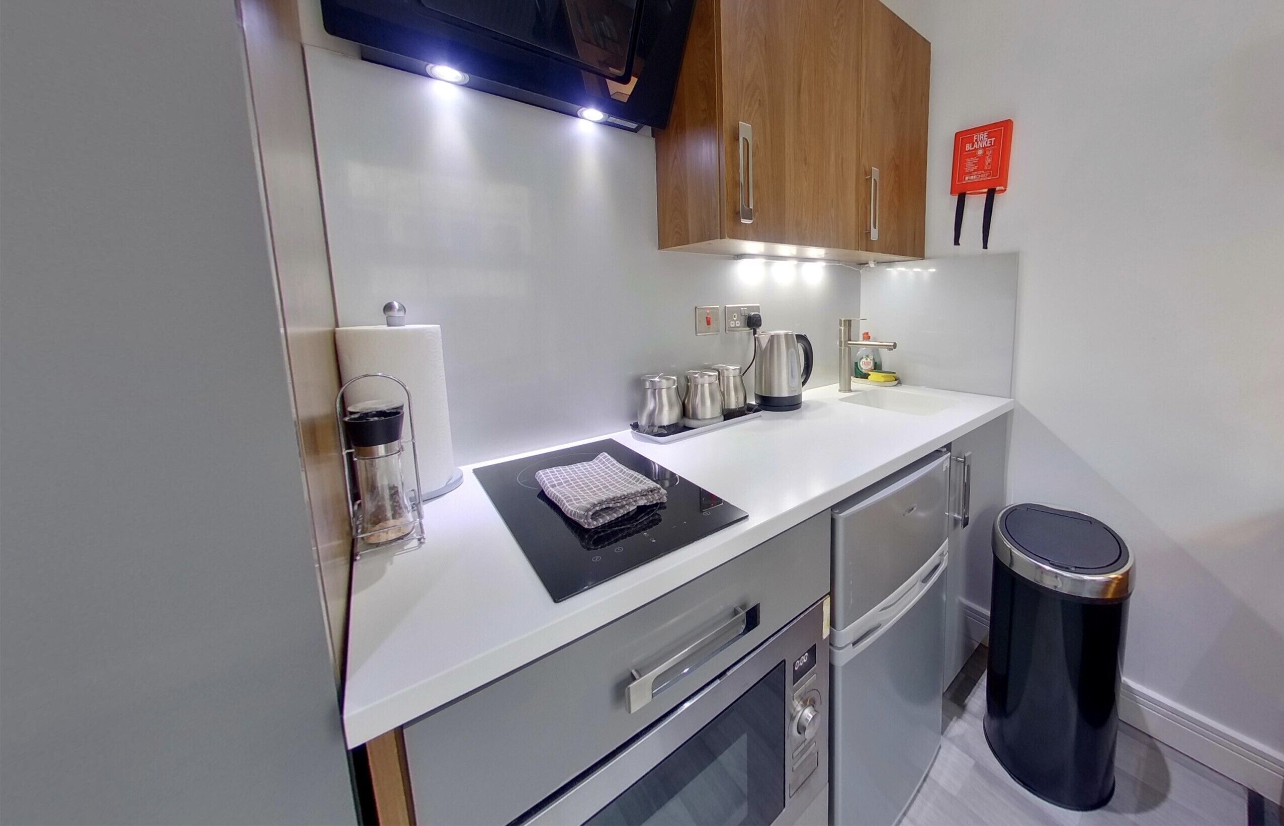 A compact kitchen with a microwave, kettle, and coffee maker on the counter, a trash bin, and wooden cabinets.