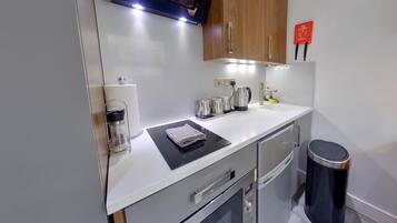 A compact kitchen with a microwave, kettle, and coffee maker on the counter, a trash bin, and wooden cabinets.