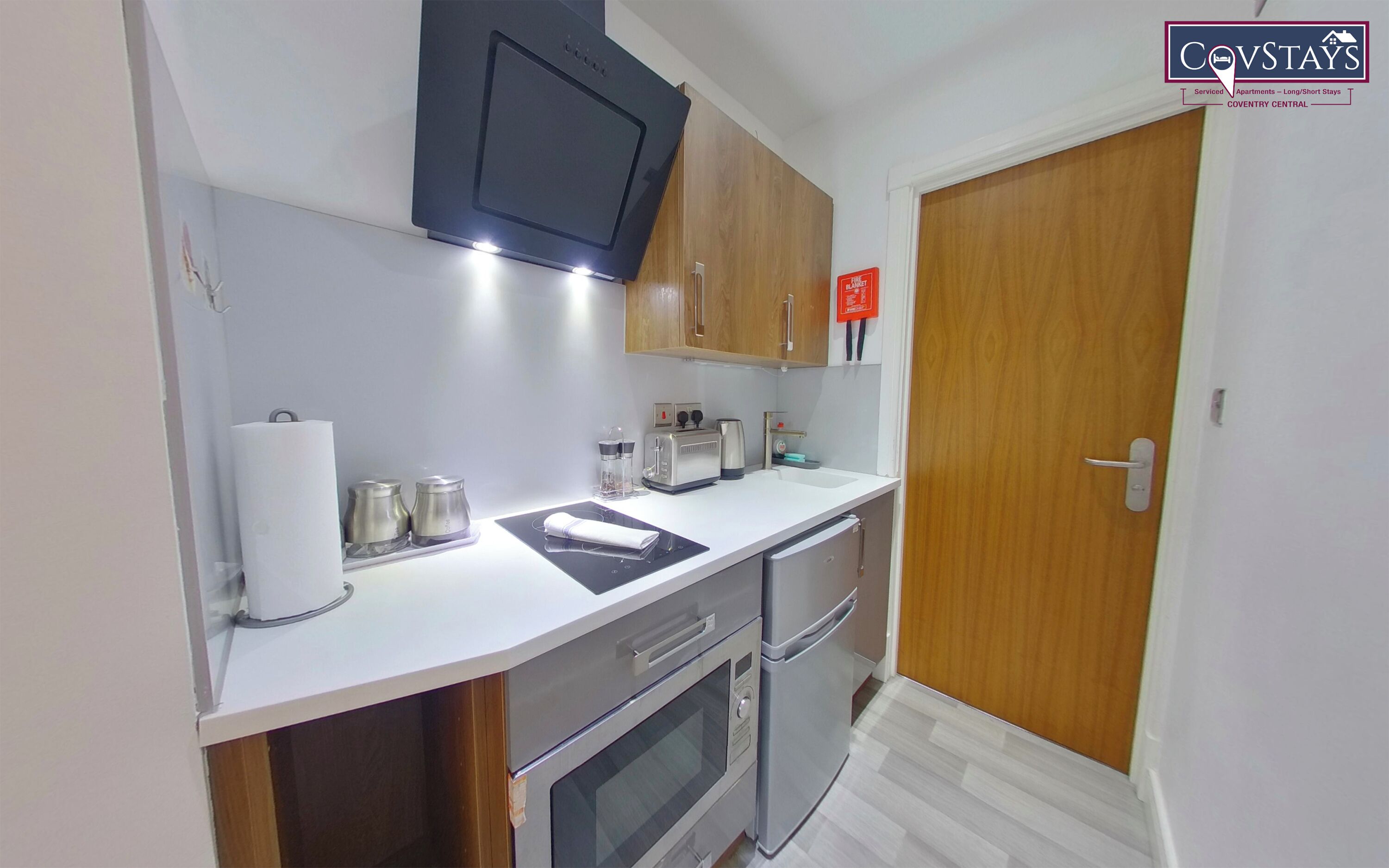 A compact kitchen with a microwave, toaster, and oven. There is a wooden door and a small shelf with items on it.
