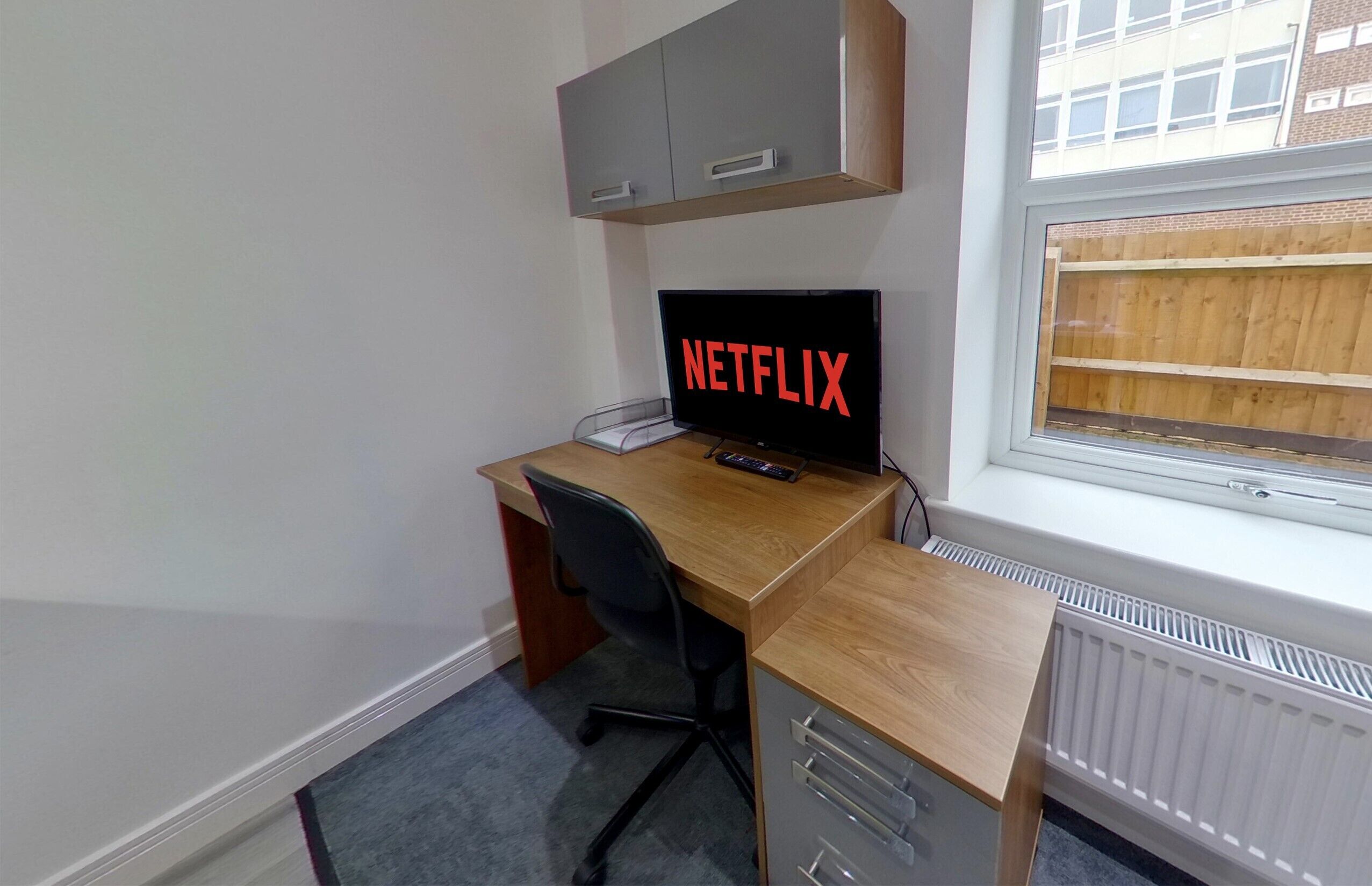 Ein kleines, ordentliches Zimmer mit einem Holztisch, einem schwarzen Bürostuhl, einem Fernseher mit dem Netflix-Logo, einem grauen Schrank über dem Tisch und einem Fenster mit Blick auf ein Gebäude.