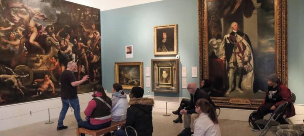 A museum interior with large paintings, a seated audience, and a guide.